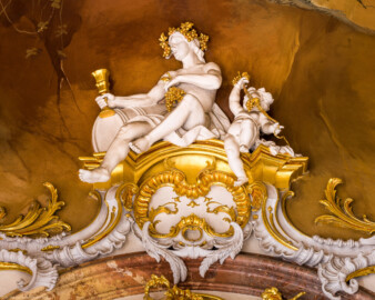 Schloss Bruchsal - Hauptbau / Corps de Logis - Beletage - Marmorsaal / Kaisersaal - Stuckaturzone, oberhalb des Durchgangs zum Jagdzimmer - Blick auf Figuren, einen Wein trinkenden Mann und einen Putto mit Weinreben (Deckenbild ursprünglich von Johannes Zick, rekonstruiert von Wolfram Köberl, 1969 bis 1973; Stuckaturen ursprünglich von Johann Michael Feichtmayr) (aufgenommen im März 2023, am Nachmittag)