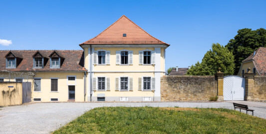 Bad Schönborn / Bad Mingolsheim - Schloss Kislau - Kavalierbau / Stallungsgebäude / Tor zum Ehrenhof - Von Süden - Blick vom Ehrenhof auf die Südfassade des Kavalierbaus (Mitte), ein ehemaliges Stallungsgebäude (links), auf die Trennmauer und das Tor zum Ehrenhof (rechts) sowie auf ein Ökonomiegebäude (ganz rechts, im Anschnitt) (aufgenommen im Juni 2023, am frühen Nachmittag)