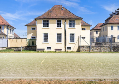Bad Schönborn / Bad Mingolsheim - Schloss Kislau - Krankenbau / Kavalierbau / Hauptbau - Von Westen - Blick vom ehemaligen Garten (heute Sportplatz) auf die Westfassade des Krankenbaus (ehemals Heiduckenbau bzw. Lakaienspeisehaus, in der Mitte), links der Kavalierbau, rechts die beiden Flügel des Hauptbaus (aufgenommen im Juni 2023, am Nachmittag)
