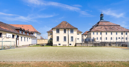 Bad Schönborn / Bad Mingolsheim - Schloss Kislau - Krankenbau / Kavalierbau / Hauptbau - Von Nordwest - Blick vom ehemaligen Garten (heute Sportplatz) auf die Westfassade des Krankenbaus (ehemals Heiduckenbau bzw. Lakaienspeisehaus, in der Mitte), links hinten der Kavalierbau, ganz links ein Stallungsgebäude,  rechts die Westfassade des Hauptbaus mit den beiden Flügeln (aufgenommen im Juni 2023, am Nachmittag)