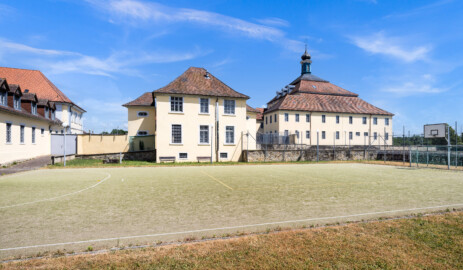 Bad Schönborn / Bad Mingolsheim - Schloss Kislau - Krankenbau / Kavalierbau / Hauptbau - Von Nordwest - Blick vom ehemaligen Garten (heute Sportplatz) auf die Westfassade des Krankenbaus (ehemals Heiduckenbau bzw. Lakaienspeisehaus, in der Mitte), links hinten der Kavalierbau, ganz links ein Stallungsgebäude,  rechts die Westfassade des Hauptbaus mit den beiden Flügeln (aufgenommen im Juni 2023, am Nachmittag)