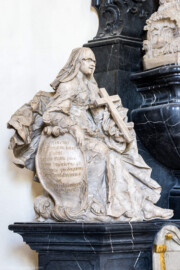 Bruchsal - Peterskirche / St. Peter - Westlicher Chorraum - Doppelgrabdenkmal für Damian Hugo Philipp von Schönborn-Buchheim und für August von Limburg-Stirum -  Von Nordost - Blick auf die Plastik / Alabasterfigur "Der Glaube" mit einem Kreuz; die beiden allegorischen Figuren "Glaube" (links) und "Liebe" (rechts) halten Schilde, auf denen eine zusammengehörende lateinische Inschrift eingehauen ist; sie lautet auf Deutsch: "Dem Verstorbenen errichtete dieses Denkmal, des leiblichen Bruders fromme Seele mit Sehnsucht und Tränen geleitend der erhabene und vornehme Herr, Herr Rudolf Franz Erwin Graf von Schönborn, Buchheim und Wollschal etc., Ritter des Ordens vom Goldenen Vlies und engster Berater ihrer kaiserlichen Majestät etc."  (Mitte links am Grabdenkmal; Skulptur gefertigt von Ferdindanz Diez) (aufgenommen im Mai 2024, am frühen Nachmittag)
