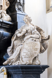 Bruchsal - Peterskirche / St. Peter - Westlicher Chorraum - Doppelgrabdenkmal für Damian Hugo Philipp von Schönborn-Buchheim und für August von Limburg-Stirum -  Von Nordost - Blick auf die Plastik / Alabasterfigur "Die Liebe" mit einem flammenden Herzen; die beiden allegorischen Figuren "Glaube" (links) und "Liebe" (rechts) halten Schilde, auf denen eine zusammengehörende lateinische Inschrift eingehauen ist; sie lautet auf Deutsch: "Dem Verstorbenen errichtete dieses Denkmal, des leiblichen Bruders fromme Seele mit Sehnsucht und Tränen geleitend der erhabene und vornehme Herr, Herr Rudolf Franz Erwin Graf von Schönborn, Buchheim und Wollschal etc., Ritter des Ordens vom Goldenen Vlies und engster Berater ihrer kaiserlichen Majestät etc." (Mitte rechts am Grabdenkmal; Skulptur gefertigt von Ferdindanz Diez) (aufgenommen im Mai 2024, am frühen Nachmittag)