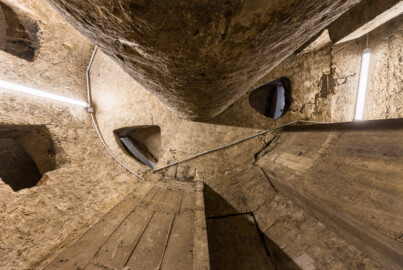 Bad Schönborn / Bad Mingolsheim - Schloss Kislau - Hauptbau - Von Innen - Treppenhaus, Keller - Blick senkrecht nach oben in den äußeren Zylinder (reicht vom Boden bis zur Decke), der das Treppenauge (der innere Zylinder vom Boden bis zur Decke, um den sich die Treppe windet) umgibt; die dreieckigen Öffnungen an der Decke (nahe der Bildmitte) sowie die rechteckigen fensterartigen Öffnungen (links im Bild) dienen der "Beleuchtung" des Kellers (aufgenommen im Mai 2024, am frühen Nachmittag)