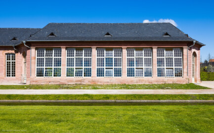 Schloss und Schlossgarten Schwetzingen - Garten - Neue Orangerie - Südseite, Glashaus - Blick vom Orangeriegarten auf die Südfassade des nach Süden hervortretenden Glashauses, mit den fünf großen schräg eingebauten Fenstern nach Süden und den drei großen Türen nach Südwesten, Südosten und Osten (Gesamter Bau: Länge ca. 171 Meter, Breite ca. 10-12 Meter; Bauzeit: 1761-1762, Glashaus angebaut 1770; Architekt: Nicolas de Pigage; umfangreiche Instandsetzung: 1994-1999; Putzflächen bemalt in Freskotechnik, täuschen teilweise ein Scheinmauerwerk vor) (aufgenommen im April 2023, um die Mittagszeit)