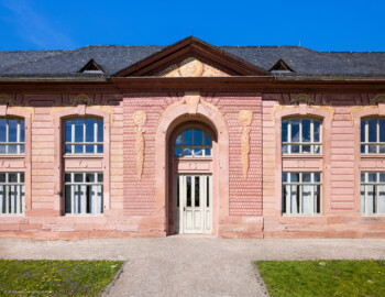 Schloss und Schlossgarten Schwetzingen - Garten - Neue Orangerie - Südseite, Mittelbau - Blick auf den fünfachsigen zweigeschossigen Mittelbau, das rundbogige Portal, den Dreiecksgiebel mit dem Sonnengesicht und den zwei Füllhörnern, die drei Löwenmasken mit Fruchtgehängen sowie die Muscheln mit Füllhörnern über den Fensterbögen (Gesamter Bau: Länge ca. 171 Meter, Breite ca. 10-12 Meter; Bauzeit: 1761-1762; Architekt: Nicolas de Pigage; umfangreiche Instandsetzung: 1994-1999; Putzflächen bemalt in Freskotechnik, täuschen teilweise ein Scheinmauerwerk vor) (aufgenommen im April 2023, am Nachmittag)