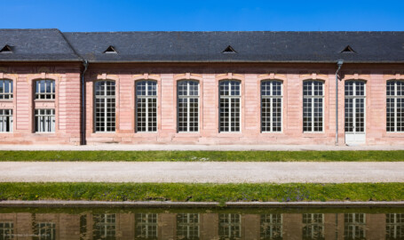 Schloss und Schlossgarten Schwetzingen - Garten - Neue Orangerie - Südseite, rechts (östlich) des Portals des Mittelbaus - Blick vom Orangeriegarten auf den westlichen Teil des östlichen dreizehnachsigen Seitenflügels mit der Tür in der Mitte des Flügels (Gesamter Bau: Länge ca. 171 Meter, Breite ca. 10-12 Meter; Bauzeit: 1761-1762; Architekt: Nicolas de Pigage; umfangreiche Instandsetzung: 1994-1999; Putzflächen bemalt in Freskotechnik, täuschen teilweise ein Scheinmauerwerk vor) (aufgenommen im April 2023, am Nachmittag) Schloss und Schlossgarten Schwetzingen - Garten - Neue Orangerie - Südseite, rechts (östlich) des Portals des Mittelbaus - Blick vom Orangeriegarten auf den westlichen Teil des östlichen dreizehnachsigen Seitenflügels mit der Tür in der Mitte des Flügels (Gesamter Bau: Länge ca. 171 Meter, Breite ca. 10-12 Meter; Bauzeit: 1761-1762; Architekt: Nicolas de Pigage; umfangreiche Instandsetzung: 1994-1999; Putzflächen bemalt in Freskotechnik, täuschen teilweise ein Scheinmauerwerk vor) (aufgenommen im April 2023, am Nachmittag)