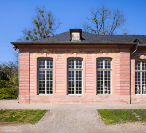 Schloss und Schlossgarten Schwetzingen - Garten - Neue Orangerie - Südseite, westlicher Eckpavillon - Blick auf die Südfassade des westlichen dreiachsigen Eckpavillons mit hohen Fenstern; dahinter lagerten früher einheimische und exotische Pflanzen; heute befindet sich dahinter ein Teil des Lapidariums mit Originalen von Skulpturen aus dem Schlossgarten (Gesamter Bau: Länge ca. 171 Meter, Breite ca. 10-12 Meter; Bauzeit: 1761-1762; Architekt: Nicolas de Pigage; umfangreiche Instandsetzung: 1994-1999; Putzflächen bemalt in Freskotechnik, täuschen teilweise ein Scheinmauerwerk vor) (aufgenommen im April 2023, am Nachmittag)