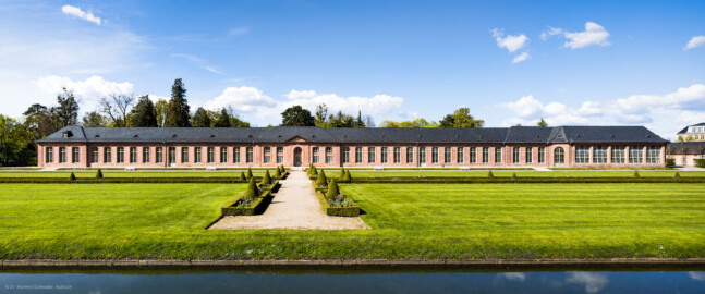 Schloss und Schlossgarten Schwetzingen - Garten - Neue Orangerie - Südseite - Blick vom Weg oberhalb des Orangeriegartens nach Norden auf die Neue Orangerie; Aufbau der Orangerie: fünfachsiger Mittelbau, zweigeschossig, mit rundbogigem Portal; links und rechts davon dreizehnachsige Seitenflügel, jeweils in der Mitte eine Tür; links und rechts der Seitenflügel dreiachsige Eckpavillons; ganz rechts ein nachträglich angebautes Glashaus; ein geplantes Glashaus ganz links ist nicht realisiert (Gesamter Bau: Länge ca. 171 Meter, Breite ca. 10-12 Meter; Bauzeit: 1761-1762, Glashaus angebaut 1770; Architekt: Nicolas de Pigage; umfangreiche Instandsetzung: 1994-1999) (aufgenommen im April 2023, am späten Nachmittag)