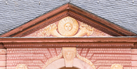 Schloss und Schlossgarten Schwetzingen - Garten - Neue Orangerie - Südseite, Mittelbau, Giebel - Blick auf den Dreiecksgiebel mit dem Sonnengesicht und den zwei Füllhörnern, darunter die drei Löwenmasken (Gesamter Bau: Länge ca. 171 Meter, Breite ca. 10-12 Meter; Bauzeit: 1761-1762; Architekt: Nicolas de Pigage; umfangreiche Instandsetzung: 1994-1999; Putzflächen bemalt in Freskotechnik, täuschen teilweise ein Scheinmauerwerk vor) (aufgenommen im Mai 2023, am späten Vormittag) Schloss und Schlossgarten Schwetzingen - Garten - Neue Orangerie - Südseite, Mittelbau, Giebel - Blick auf den Dreiecksgiebel mit dem Sonnengesicht und den zwei Füllhörnern, darunter die drei Löwenmasken (Gesamter Bau: Länge ca. 171 Meter, Breite ca. 10-12 Meter; Bauzeit: 1761-1762; Architekt: Nicolas de Pigage; umfangreiche Instandsetzung: 1994-1999; Putzflächen bemalt in Freskotechnik, täuschen teilweise ein Scheinmauerwerk vor) (aufgenommen im Mai 2023, am späten Vormittag)