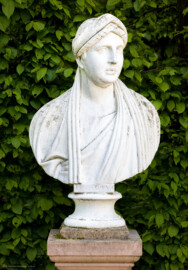 Schloss und Schlossgarten Schwetzingen - Garten - Porzellanhaus, Rasenfläche südlich, Südwestecke - Büste von Faustina der Jüngeren (Annia Galeria Faustina, 130-176 n.Chr., Ehefrau des römischen Kaisers Mark Aurel, 121-180 n.Chr.); gehauen von Franz Conrad Linck (1730-1793) nach einer antiken Vorlage aus dem Mannheimer Antikensaal; Büste aus Sandstein, weiß gefaßt, um Marmor vorzutäuschen, auf einem Sandsteinpostament stehend; die Büste gehört zu einer Sechsergruppe (Faustina, Domitian, Marciana, Marcellus, Solon, Gladiator) (aufgenommen im Mai 2023, am Nachmittag)