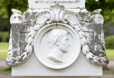 Schloss und Schlossgarten Schwetzingen - Garten - Kreisparterre, nordwestliches Rasenfeld - Nordwestlicher Obelisk - Von Osten - Blick auf ein männnliches Profilbildnis / Flachrelief auf der Ostseite des Obelisken (Person nicht eindeutig identifizierbar) (Bildhauer: Peter Anton von Verschaffelt (1710-1793); Datierung: 1762-69; Obelisk Teil einer Vierergruppe im Kreisparterre; Material: roter Sandstein (Medaillons aus gelbem Sandstein); Fassung: Weiß; Obelisk dreigeteilt; Sockel, Block mit Medaillonbildnissen, eigentlicher Obelisk; Abmessungen: Höhe ca. 6 Meter, Sockel Höhe 155 cm / Breite 87 cm, Block mit Medaillons Höhe 67 cm / Breite 87 cm; Obelisk oben mit Tropfsteindekor) (aufgenommen im Mai 2023, am späten Nachmittag)