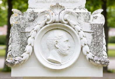 Schloss und Schlossgarten Schwetzingen - Garten - Kreisparterre, nordwestliches Rasenfeld- Nordwestlicher Obelisk - Von Westen - Blick auf ein männnliches Profilbildnis / Flachrelief auf der Westseite des Obelisken (Person nicht eindeutig identifizierbar) (Bildhauer: Peter Anton von Verschaffelt (1710-1793); Datierung: 1762-69; Obelisk Teil einer Vierergruppe im Kreisparterre; Material: roter Sandstein (Medaillons aus gelbem Sandstein); Fassung: Weiß; Obelisk dreigeteilt; Sockel, Block mit Medaillonbildnissen, eigentlicher Obelisk; Abmessungen: Höhe ca. 6 Meter, Sockel Höhe 155 cm / Breite 87 cm, Block mit Medaillons Höhe 67 cm / Breite 87 cm; Obelisk oben mit Tropfsteindekor) (aufgenommen im Mai 2023, am späten Nachmittag)