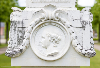 Schloss und Schlossgarten Schwetzingen - Garten - Kreisparterre, nordwestliches Rasenfeld - Nordwestlicher Obelisk - Von Süden - Blick auf ein weibliches Profilbildnis / Flachrelief auf der Südseite des Obelisken (Person nicht eindeutig identifizierbar) (Bildhauer: Peter Anton von Verschaffelt (1710-1793); Datierung: 1762-69; Obelisk Teil einer Vierergruppe im Kreisparterre; Material: roter Sandstein (Medaillons aus gelbem Sandstein); Fassung: Weiß; Obelisk dreigeteilt; Sockel, Block mit Medaillonbildnissen, eigentlicher Obelisk; Abmessungen: Höhe ca. 6 Meter, Sockel Höhe 155 cm / Breite 87 cm, Block mit Medaillons Höhe 67 cm / Breite 87 cm; Obelisk oben mit Tropfsteindekor) (aufgenommen im Mai 2023, am späten Nachmittag)