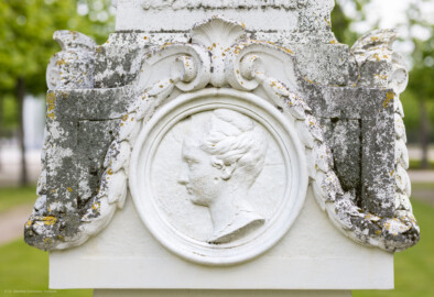Schloss und Schlossgarten Schwetzingen - Garten - Kreisparterre, nordwestliches Rasenfeld - Nordwestlicher Obelisk - Von Norden - Blick auf ein weibliches Profilbildnis / Flachrelief auf der Nordseite des Obelisken (Person nicht eindeutig identifizierbar) (Bildhauer: Peter Anton von Verschaffelt (1710-1793); Datierung: 1762-69; Obelisk Teil einer Vierergruppe im Kreisparterre; Material: roter Sandstein (Medaillons aus gelbem Sandstein); Fassung: Weiß; Obelisk dreigeteilt; Sockel, Block mit Medaillonbildnissen, eigentlicher Obelisk; Abmessungen: Höhe ca. 6 Meter, Sockel Höhe 155 cm / Breite 87 cm, Block mit Medaillons Höhe 67 cm / Breite 87 cm; Obelisk oben mit Tropfsteindekor) (aufgenommen im Mai 2023, am späten Nachmittag)