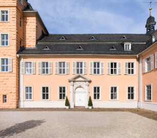 Schloss und Schlossgarten Schwetzingen - Schloss - Nordflügel / Hofkavaliersflügel - Querbau - Von Osten - Blick auf die Schlosskapelle mit dem Portal, ganz links der Nordturm, oben rechts daneben ein Teil des Mittelbaus, oben rechts der Dachreiter des Nordflügels mit einem Hemmerschen Fünfspitz und ganz rechts der Längsbau des Nordflügels; im oberen linken Fenster (in Höhe der Hofloge) der Schlosskapelle blickt Carl Theodor (eine gemalte Figur) aus dem (gemalten) Fenster auf den Ehrenhof herunter; das rechte obere Fenster der Schlosskapelle gehört zur Orgelempore, das Fenster darunter zur Sakristei (NordFlügel: L-förmig, Länge x Breite Längsbau ca. 50 x 14 Meter, Querbau ca. 15 x 12 Meter; Architekt: Johann Adam Breunig (1660-1727); Fertigstellung: 1710) (aufgenommen im Mai 2023, am Vormittag)
