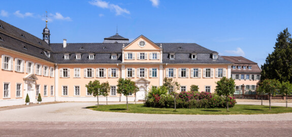 Schloss und Schlossgarten Schwetzingen - Schloss - Nordflügel / Hofkavaliersflügel - Von Süden - Blick vom Ehrenhof nach Norden auf den Ehrenhof, auf die Südfassade des Längsbaus (Mitte; heute genutzt vom Finanzamt) sowie auf die Ostfassade des Querbaus bzw. der Schlosskapelle (links); links der Mitte oben im Hintergrund das Dach des Oberen Wasserwerks (NordFlügel: L-förmig, Länge x Breite Längsbau ca. 50 x 14 Meter, Querbau ca. 15 x 12 Meter; Architekt: Johann Adam Breunig (1660-1727); Fertigstellung: 1710) (aufgenommen im Mai 2023, um die Mittagszeit)