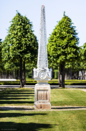 Schloss und Schlossgarten Schwetzingen - Garten - Kreisparterre, südostliches Rasenfeld - Südostlicher Obelisk - Von Süden - Blick auf die Südseite des Obelisken (Bildhauer: Peter Anton von Verschaffelt (1710-1793); Datierung: 1762-69; Obelisk Teil einer Vierergruppe im Kreisparterre; Material: roter Sandstein (Medaillons aus gelbem Sandstein); Fassung: Weiß; Obelisk dreigeteilt; Sockel, Block mit Medaillonbildnissen, eigentlicher Obelisk; Abmessungen: Höhe ca. 6 Meter, Sockel Höhe 155 cm / Breite 87 cm, Block mit Medaillons Höhe 67 cm / Breite 87 cm; Obelisk oben mit Tropfsteindekor) (aufgenommen im Mai 2023, am Nachmittag)