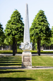 Schloss und Schlossgarten Schwetzingen - Garten - Kreisparterre, südwestliches Rasenfeld - Südwestlicher Obelisk - Von Süden - Blick auf die Südseite des Obelisken (Bildhauer: Peter Anton von Verschaffelt (1710-1793); Datierung: 1762-69; Obelisk Teil einer Vierergruppe im Kreisparterre; Material: roter Sandstein (Medaillons aus gelbem Sandstein); Fassung: Weiß; Obelisk dreigeteilt; Sockel, Block mit Medaillonbildnissen, eigentlicher Obelisk; Abmessungen: Höhe ca. 6 Meter, Sockel Höhe 155 cm / Breite 87 cm, Block mit Medaillons Höhe 67 cm / Breite 87 cm; Obelisk oben mit Tropfsteindekor) (aufgenommen im Mai 2023, am Nachmittag)