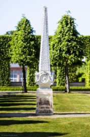 Schloss und Schlossgarten Schwetzingen - Garten - Kreisparterre, nordwestliches Rasenfeld - Nordwestlicher Obelisk - Von Süden - Blick auf die Südseite des Obelisken (Bildhauer: Peter Anton von Verschaffelt (1710-1793); Datierung: 1762-69; Obelisk Teil einer Vierergruppe im Kreisparterre; Material: roter Sandstein (Medaillons aus gelbem Sandstein); Fassung: Weiß; Obelisk dreigeteilt; Sockel, Block mit Medaillonbildnissen, eigentlicher Obelisk; Abmessungen: Höhe ca. 6 Meter, Sockel Höhe 155 cm / Breite 87 cm, Block mit Medaillons Höhe 67 cm / Breite 87 cm; Obelisk oben mit Tropfsteindekor) (aufgenommen im Mai 2023, am späten Nachmittag)