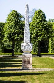 Schloss und Schlossgarten Schwetzingen - Garten - Kreisparterre, nordöstliches Rasenfeld - Nordöstlicher Obelisk - Von Süden - Blick auf die Südseite des Obelisken (Bildhauer: Peter Anton von Verschaffelt (1710-1793); Datierung: 1762-69; Obelisk Teil einer Vierergruppe im Kreisparterre; Material: roter Sandstein (Medaillons aus gelbem Sandstein); Fassung: Weiß; Obelisk dreigeteilt; Sockel, Block mit Medaillonbildnissen, eigentlicher Obelisk; Abmessungen: Höhe ca. 6 Meter, Sockel Höhe 155 cm / Breite 87 cm, Block mit Medaillons Höhe 67 cm / Breite 87 cm; Obelisk oben mit Tropfsteindekor) (aufgenommen im Mai 2023, am späten Nachmittag)