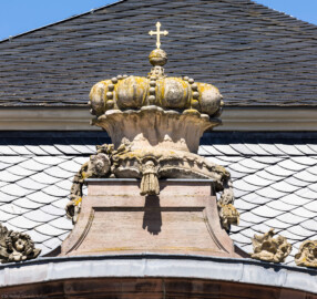 Schloss und Schlossgarten Schwetzingen - Garten - Nördlicher Zirkelbau - Östlicher Eck-Pavillon - Südportal - Von Süden - Blick nach oben auf die Krone mit Kreuz über dem Südportal; Krone ist eine Kopie, Original der Skulptur vermutlich gehauen von Johann Matthäus van den Branden (1716-1788) (Gesamter Bau: Länge Bogen ca. 175 Meter, Breite zwischen ca. 11 und 18 Meter, 51 Achsen, 5 Pavillons, 4 Zwischenbauten; Architekt: Alessandro Alessandro Galli da Bibiena (1686-1748), Bauleitung: zuerst Guillaume d’Hauberat (1680–1749), dann Nicolas de Pigage (1723-1796); Bauzeit: 1748-1750) (aufgenommen im August 2023, am frühen Nachmittag)