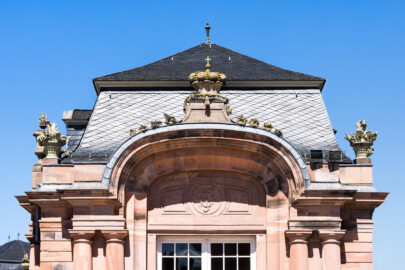 Schloss und Schlossgarten Schwetzingen - Garten - Nördlicher Zirkelbau - Östlicher Eck-Pavillon - Südportal - Von Süden - Blick nach oben auf das Südportal und das Dach des Pavillon, auf das Monogramm "CT" (Carl Theodor) im Bogen, auf die Krone mit Kreuz sowie auf Blumenkörbe mit Blumen und Vasen mit Flammen; Krone, Blumenkörbe und Vasen sind Kopien, Originale der Skulpturen vermutlich gehauen von Johann Matthäus van den Branden (1716-1788) (Gesamter Bau: Länge Bogen ca. 175 Meter, Breite zwischen ca. 11 und 18 Meter, 51 Achsen, 5 Pavillons, 4 Zwischenbauten; Architekt: Alessandro Alessandro Galli da Bibiena (1686-1748), Bauleitung: zuerst Guillaume d’Hauberat (1680–1749), dann Nicolas de Pigage (1723-1796); Bauzeit: 1748-1750) (aufgenommen im August 2023, am frühen Nachmittag)