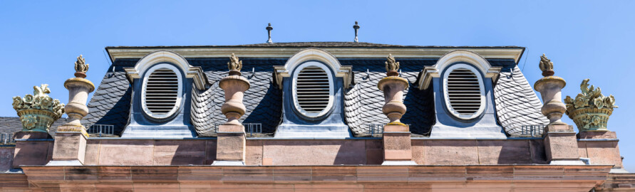 Schloss und Schlossgarten Schwetzingen - Garten - Nördlicher Zirkelbau - Östlicher Eck-Pavillon - Von Westen - Blick nach oben auf das Dach des Pavillons, die drei Ovalfenster, die vier Vasen mit Flammen sowie die zwei Blumenkörbe mit Blumen; Vasen und Blumenkörbe sind Kopien, Originale der Skulpturen vermutlich gehauen von Johann Matthäus van den Branden (1716-1788) (Gesamter Bau: Länge Bogen ca. 175 Meter, Breite zwischen ca. 11 und 18 Meter, 51 Achsen, 5 Pavillons, 4 Zwischenbauten; Architekt: Alessandro Alessandro Galli da Bibiena (1686-1748), Bauleitung: zuerst Guillaume d’Hauberat (1680–1749), dann Nicolas de Pigage (1723-1796); Bauzeit: 1748-1750) (aufgenommen im August 2023, am frühen Nachmittag)