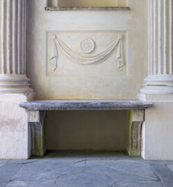 Schloss und Schlossgarten Schwetzingen - Garten - Minervatempel - Von innen - Linke Seitenwand, zwischen vorderem Säulenpaar - Blick auf ein etwas vertieftes Feld mit einem in Stuck ausgeführtes Relief, ein Tuchgehänge mit einer Rosette sowie auf eine Steinbank zwischen den Postamenten der Säulen (Gesamter Bau: Breite ca. 7 Meter, Länge ca. 9 Meter, Innenraum kubisch; Rückseite zweigeschossig mit Kellerraum; Architekt: Nicolas de Pigage (1723-1796); Bauzeit: vermutlich zwischen 1767/68 und 1773) (aufgenommen im August 2023, am späten Vormittag)