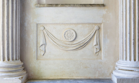 Schloss und Schlossgarten Schwetzingen - Garten - Minervatempel - Von innen - Linke Seitenwand, zwischen vorderem Säulenpaar - Blick auf ein etwas vertieftes Feld mit einem in Stuck ausgeführtes Relief, ein Tuchgehänge mit einer Rosette (Gesamter Bau: Breite ca. 7 Meter, Länge ca. 9 Meter, Innenraum kubisch; Rückseite zweigeschossig mit Kellerraum; Architekt: Nicolas de Pigage (1723-1796); Bauzeit: vermutlich zwischen 1767/68 und 1773) (aufgenommen im August 2023, am späten Vormittag)