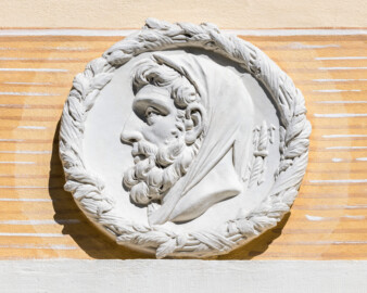 Schloss und Schlossgarten Schwetzingen - Garten - Badhaus - Von aussen - Westseite - Medaillon oben rechts - Von Westen - Blick auf ein Feld mit einem Relief-Medaillon; dargestellt ist der Kopf von Zeus, links gewendet, das Lockenhaupt mit einem Tuch bedeckt, am Hinterkopf zwei stilisierte Doppelblitze (Bildhauer: Franz Conrad Linck (1730-1793), Datierung: 1770-1773; Kopie, Verbleib Original unklar; Material: Terrakotta; Durchmesser: ca. 40cm) (Gesamter Bau: Länge x Breite ca. 18 x 14 Meter, Höhe ca. 10 Meter; Architekt: Nicolas de Pigage (1723-1796); Bauzeit: 1768-1772; Gesamtinstandsetzung innen und außen: 1999-2006) (aufgenommen im August 2023, am späten Nachmittag)