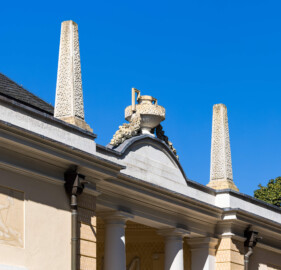 Schloss und Schlossgarten Schwetzingen - Garten - Badhaus - Von aussen - Südseite - Obelisken, oberhalb Eingangsportal - Von Südwest - Blick nach oben auf die beiden Obelisken oberhalb des südlichen Eingangs; Obelisken mit geböschtem Sockel, sehr flacher Spitze, mit Tropfen aus Stuck; in der Mitte eine bauchige Vase im antikisierenden Stil mit rechteckigen Henkeln und Fruchtgirlanden (Bildhauer der Vase: Franz Conrad Linck (1730-1793)) (Gesamter Bau: Länge x Breite ca. 18 x 14 Meter, Höhe ca. 10 Meter; Architekt: Nicolas de Pigage (1723-1796); Bauzeit: 1768-1772; Gesamtinstandsetzung innen und außen: 1999-2006) (aufgenommen im August 2023, am späten Nachmittag)