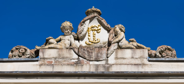 Schloss und Schlossgarten Schwetzingen - Garten - Badhaus - Von aussen - Westseite - Kartusche mit Monogramm oberhalb Mittelrisalit - Von Westen - Blick nach oben auf eine ovale Kartusche mit den verschlungenen, vergoldeten Initialen / dem Monogramm von Carl Theodor ("CT"); rechts und links zwei Putti und außen zwei Füllhörner, mit Früchten gefüllt, darüber der Kurhut mit Kreuz (Bildhauer: Franz Conrad Linck (1730-1793), Datierung: 1770; Material: Sandstein) (Gesamter Bau: Länge x Breite ca. 18 x 15 Meter, Höhe ca. 11 Meter; Architekt: Nicolas de Pigage (1723-1796); Bauzeit: 1768-1772; Gesamtinstandsetzung innen und außen: 1999-2006) (aufgenommen im August 2023, am späten Nachmittag) Schloss und Schlossgarten Schwetzingen - Garten - Badhaus - Von aussen - Westseite - Kartusche mit Monogramm oberhalb Mittelrisalit - Von Westen - Blick nach oben auf eine ovale Kartusche mit den verschlungenen, vergoldeten Initialen / dem Monogramm von Carl Theodor ("CT"); rechts und links zwei Putti und außen zwei Füllhörner, mit Früchten gefüllt, darüber der Kurhut mit Kreuz (Bildhauer: Franz Conrad Linck (1730-1793), Datierung: 1770; Material: Sandstein) (Gesamter Bau: Länge x Breite ca. 18 x 15 Meter, Höhe ca. 11 Meter; Architekt: Nicolas de Pigage (1723-1796); Bauzeit: 1768-1772; Gesamtinstandsetzung innen und außen: 1999-2006) (aufgenommen im August 2023, am späten Nachmittag)