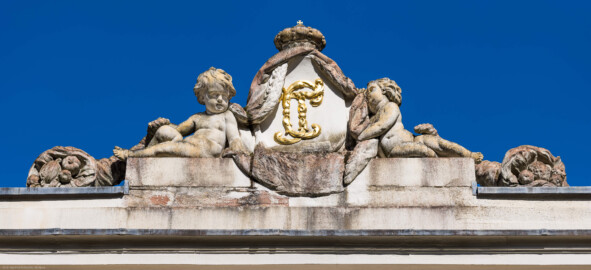 Schloss und Schlossgarten Schwetzingen - Garten - Badhaus - Von aussen - Westseite - Kartusche mit Monogramm oberhalb Mittelrisalit - Von Westen - Blick nach oben auf eine ovale Kartusche mit den verschlungenen, vergoldeten Initialen / dem Monogramm von Carl Theodor ("CT"); rechts und links zwei Putti und außen zwei Füllhörner, mit Früchten gefüllt, darüber der Kurhut mit Kreuz (Bildhauer: Franz Conrad Linck (1730-1793), Datierung: 1770; Material: Sandstein) (Gesamter Bau: Länge x Breite ca. 18 x 14 Meter, Höhe ca. 10 Meter; Architekt: Nicolas de Pigage (1723-1796); Bauzeit: 1768-1772; Gesamtinstandsetzung innen und außen: 1999-2006) (aufgenommen im August 2023, am späten Nachmittag)
