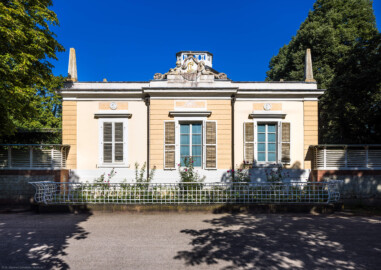 Schloss und Schlossgarten Schwetzingen - Garten - Badhaus - Von aussen - Westseite - Von Westen - Blick auf die Westseite des Badhauses, gegliedert mit drei Achsen, Ecklisenen und mittigem Risalit; in der Mitte oben ein Feld mit Jakobsmuschel und Lorbeerkranz, links oben ein Feld mit Alexander-Medaillon, rechts oben mit Zeus-Medaillon, mittig ganz oben eine ovale Kartusche, von zwei Putti gehalten, mit den Initialen von Carl Theodor ("CT"), darüber der Kurhut mit Kreuz, dahinter das Geländer des Tambours, auf den Ecken oben zwei der acht Obelisken (Gesamter Bau: Länge x Breite ca. 18 x 14 Meter, Höhe ca. 10 Meter; Architekt: Nicolas de Pigage (1723-1796); Bauzeit: 1768-1772; Gesamtinstandsetzung innen und außen: 1999-2006) (aufgenommen im August 2023, am späten Nachmittag)
