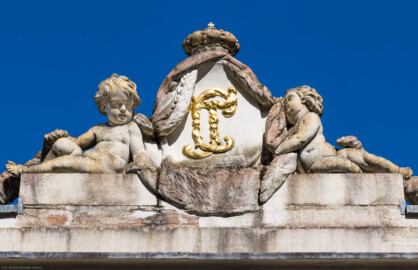 Schloss und Schlossgarten Schwetzingen - Garten - Badhaus - Von aussen - Westseite - Kartusche mit Monogramm oberhalb Mittelrisalit - Von Westen - Blick nach oben auf eine ovale Kartusche mit den verschlungenen, vergoldeten Initialen / dem Monogramm von Carl Theodor ("CT"); rechts und links zwei Putti und außen zwei Füllhörner, mit Früchten gefüllt, darüber der Kurhut mit Kreuz (Bildhauer: Franz Conrad Linck (1730-1793), Datierung: 1770; Material: Sandstein) (Gesamter Bau: Länge x Breite ca. 18 x 14 Meter, Höhe ca. 10 Meter; Architekt: Nicolas de Pigage (1723-1796); Bauzeit: 1768-1772; Gesamtinstandsetzung innen und außen: 1999-2006) (aufgenommen im August 2023, am späten Nachmittag)