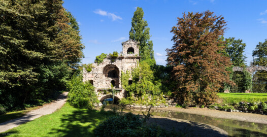 Schloss und Schlossgarten Schwetzingen - Garten - Römisches Wasserkastell - Von außen - Von Süden - Blick auf das Römische Wasserkastell; eine künstliche Ruine, teils in Tuffstein, mit zwei Türmen (westlich zwei-, östlich dreigeschossig), Mittelpartie mit großem Rundbogen, darüber eine Plattform, darunter ein Gewölbe mit Durchfluss des Leimbachs nach Norden; im östlichen Mittelgeschoss ein Tonrelief mit einer Wassernymphe von Franz Conrad Linck (1730-1793); nach Westen, Osten und Norden Aquädukte, ostseitig mit 22 Pfeilern, westseitig mit 4 Pfeilern, Nordteil mit Verbindung zum Unteren Wasserwerk (Kastell: Länge x Breite: ca. 15 x 8 Meter; Bauzeit: 1779-1780; Architekt: Nicolas de Pigage) (aufgenommen im September 2023, am späten Vormittag)