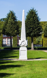 Schloss und Schlossgarten Schwetzingen - Garten - Kreisparterre, nordwestliches Rasenfeld - Nordwestlicher Obelisk - Von Süden - Blick auf die Südseite des Obelisken (Bildhauer: Peter Anton von Verschaffelt (1710-1793); Datierung: 1762-69; Obelisk Teil einer Vierergruppe im Kreisparterre; Material: roter Sandstein (Medaillons aus gelbem Sandstein); Fassung: Weiß; Obelisk dreigeteilt; Sockel, Block mit Medaillonbildnissen, eigentlicher Obelisk; Abmessungen: Höhe ca. 6 Meter, Sockel Höhe 155 cm / Breite 87 cm, Block mit Medaillons Höhe 67 cm / Breite 87 cm; Obelisk oben mit Tropfsteindekor) (aufgenommen im September 2023, am Nachmittag)