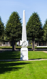 Schloss und Schlossgarten Schwetzingen - Garten - Kreisparterre, südwestliches Rasenfeld - Südwestlicher Obelisk - Von Süden - Blick auf die Südseite des Obelisken (Bildhauer: Peter Anton von Verschaffelt (1710-1793); Datierung: 1762-69; Obelisk Teil einer Vierergruppe im Kreisparterre; Material: roter Sandstein (Medaillons aus gelbem Sandstein); Fassung: Weiß; Obelisk dreigeteilt; Sockel, Block mit Medaillonbildnissen, eigentlicher Obelisk; Abmessungen: Höhe ca. 6 Meter, Sockel Höhe 155 cm / Breite 87 cm, Block mit Medaillons Höhe 67 cm / Breite 87 cm; Obelisk oben mit Tropfsteindekor) (aufgenommen im September 2023, am Nachmittag)