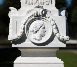 Schloss und Schlossgarten Schwetzingen - Garten - Kreisparterre, südwestliches Rasenfeld, entlang der Nord-Süd-Achse - Südwestlicher Obelisk, Südseite - Blick auf ein weibliches verwittertes Profilbildnis / Flachrelief, umgeben von einem Lorbeerfeston (Person nicht eindeutig identifizierbar) (Bildhauer: Peter Anton von Verschaffelt (1710-1793); Datierung: 1762-69; Obelisk Teil einer Vierergruppe im Kreisparterre; Material: roter Sandstein (Medaillons aus gelbem Sandstein); Fassung: Weiß; Obelisk dreigeteilt; Sockel, Block mit Medaillonbildnissen, eigentlicher Obelisk; Abmessungen: Höhe ca. 6 Meter, Sockel Höhe 155 cm / Breite 87 cm, Block mit Medaillons Höhe 67 cm / Breite 87 cm; Obelisk oben mit Tropfsteindekor) (aufgenommen im September 2023, am Nachmittag) Schloss und Schlossgarten Schwetzingen - Garten - Kreisparterre, südwestliches Rasenfeld, entlang der Nord-Süd-Achse - Südwestlicher Obelisk, Südseite - Blick auf ein weibliches verwittertes Profilbildnis / Flachrelief, umgeben von einem Lorbeerfeston (Person nicht eindeutig identifizierbar) (Bildhauer: Peter Anton von Verschaffelt (1710-1793); Datierung: 1762-69; Obelisk Teil einer Vierergruppe im Kreisparterre; Material: roter Sandstein (Medaillons aus gelbem Sandstein); Fassung: Weiß; Obelisk dreigeteilt; Sockel, Block mit Medaillonbildnissen, eigentlicher Obelisk; Abmessungen: Höhe ca. 6 Meter, Sockel Höhe 155 cm / Breite 87 cm, Block mit Medaillons Höhe 67 cm / Breite 87 cm; Obelisk oben mit Tropfsteindekor) (aufgenommen im September 2023, am Nachmittag)