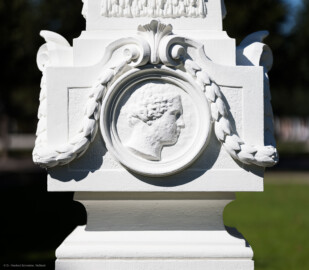 Schloss und Schlossgarten Schwetzingen - Garten - Kreisparterre, südöstliches Rasenfeld, entlang der Nord-Süd-Achse - Südöstlicher Obelisk, Südseite - Blick auf ein weibliches verwittertes Profilbildnis / Flachrelief, umgeben von einem Lorbeerfeston (Person nicht eindeutig identifizierbar) (Bildhauer: Peter Anton von Verschaffelt (1710-1793); Datierung: 1762-69; Obelisk Teil einer Vierergruppe im Kreisparterre; Material: roter Sandstein (Medaillons aus gelbem Sandstein); Fassung: Weiß; Obelisk dreigeteilt; Sockel, Block mit Medaillonbildnissen, eigentlicher Obelisk; Abmessungen: Höhe ca. 6 Meter, Sockel Höhe 155 cm / Breite 87 cm, Block mit Medaillons Höhe 67 cm / Breite 87 cm; Obelisk oben mit Tropfsteindekor) (aufgenommen im September 2023, am Nachmittag) Schloss und Schlossgarten Schwetzingen - Garten - Kreisparterre, südöstliches Rasenfeld, entlang der Nord-Süd-Achse - Südöstlicher Obelisk, Südseite - Blick auf ein weibliches verwittertes Profilbildnis / Flachrelief, umgeben von einem Lorbeerfeston (Person nicht eindeutig identifizierbar) (Bildhauer: Peter Anton von Verschaffelt (1710-1793); Datierung: 1762-69; Obelisk Teil einer Vierergruppe im Kreisparterre; Material: roter Sandstein (Medaillons aus gelbem Sandstein); Fassung: Weiß; Obelisk dreigeteilt; Sockel, Block mit Medaillonbildnissen, eigentlicher Obelisk; Abmessungen: Höhe ca. 6 Meter, Sockel Höhe 155 cm / Breite 87 cm, Block mit Medaillons Höhe 67 cm / Breite 87 cm; Obelisk oben mit Tropfsteindekor) (aufgenommen im September 2023, am Nachmittag)