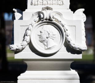 Schloss und Schlossgarten Schwetzingen - Garten - Kreisparterre, südöstliches Rasenfeld, entlang der Nord-Süd-Achse - Südöstlicher Obelisk, Westseite - Blick auf ein männliches verwittertes Profilbildnis / Flachrelief, umgeben von einem Lorbeerfeston (Person nicht eindeutig identifizierbar) (Bildhauer: Peter Anton von Verschaffelt (1710-1793); Datierung: 1762-69; Obelisk Teil einer Vierergruppe im Kreisparterre; Material: roter Sandstein (Medaillons aus gelbem Sandstein); Fassung: Weiß; Obelisk dreigeteilt; Sockel, Block mit Medaillonbildnissen, eigentlicher Obelisk; Abmessungen: Höhe ca. 6 Meter, Sockel Höhe 155 cm / Breite 87 cm, Block mit Medaillons Höhe 67 cm / Breite 87 cm; Obelisk oben mit Tropfsteindekor) (aufgenommen im September 2023, am Nachmittag)