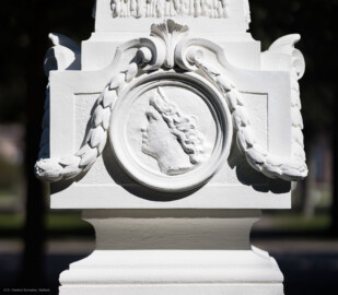 Schloss und Schlossgarten Schwetzingen - Garten - Kreisparterre, südwestliches Rasenfeld, entlang der Nord-Süd-Achse - Südwestlicher Obelisk, Westseite - Blick auf ein weibliches verwittertes Profilbildnis / Flachrelief, umgeben von einem Lorbeerfeston (Person nicht eindeutig identifizierbar) (Bildhauer: Peter Anton von Verschaffelt (1710-1793); Datierung: 1762-69; Obelisk Teil einer Vierergruppe im Kreisparterre; Material: roter Sandstein (Medaillons aus gelbem Sandstein); Fassung: Weiß; Obelisk dreigeteilt; Sockel, Block mit Medaillonbildnissen, eigentlicher Obelisk; Abmessungen: Höhe ca. 6 Meter, Sockel Höhe 155 cm / Breite 87 cm, Block mit Medaillons Höhe 67 cm / Breite 87 cm; Obelisk oben mit Tropfsteindekor) (aufgenommen im September 2023, am Nachmittag)