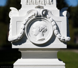 Schloss und Schlossgarten Schwetzingen - Garten - Kreisparterre, nordwestliches Rasenfeld, entlang der Nord-Süd-Achse - Nordwestlicher Obelisk, Südseite - Blick auf ein weibliches verwittertes Profilbildnis / Flachrelief, umgeben von einem Lorbeerfeston (Person nicht eindeutig identifizierbar) (Bildhauer: Peter Anton von Verschaffelt (1710-1793); Datierung: 1762-69; Obelisk Teil einer Vierergruppe im Kreisparterre; Material: roter Sandstein (Medaillons aus gelbem Sandstein); Fassung: Weiß; Obelisk dreigeteilt; Sockel, Block mit Medaillonbildnissen, eigentlicher Obelisk; Abmessungen: Höhe ca. 6 Meter, Sockel Höhe 155 cm / Breite 87 cm, Block mit Medaillons Höhe 67 cm / Breite 87 cm; Obelisk oben mit Tropfsteindekor) (aufgenommen im September 2023, am Nachmittag) Schloss und Schlossgarten Schwetzingen - Garten - Kreisparterre, nordwestliches Rasenfeld, entlang der Nord-Süd-Achse - Nordwestlicher Obelisk, Südseite - Blick auf ein weibliches verwittertes Profilbildnis / Flachrelief, umgeben von einem Lorbeerfeston (Person nicht eindeutig identifizierbar) (Bildhauer: Peter Anton von Verschaffelt (1710-1793); Datierung: 1762-69; Obelisk Teil einer Vierergruppe im Kreisparterre; Material: roter Sandstein (Medaillons aus gelbem Sandstein); Fassung: Weiß; Obelisk dreigeteilt; Sockel, Block mit Medaillonbildnissen, eigentlicher Obelisk; Abmessungen: Höhe ca. 6 Meter, Sockel Höhe 155 cm / Breite 87 cm, Block mit Medaillons Höhe 67 cm / Breite 87 cm; Obelisk oben mit Tropfsteindekor) (aufgenommen im September 2023, am Nachmittag)
