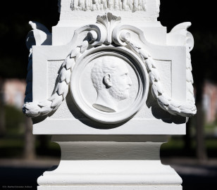 Schloss und Schlossgarten Schwetzingen - Garten - Kreisparterre, nordwestliches Rasenfeld, entlang der Nord-Süd-Achse - Nordwestlicher Obelisk, Westseite - Blick auf ein männnliches verwittertes Profilbildnis / Flachrelief, umgeben von einem Lorbeerfeston (Person nicht eindeutig identifizierbar) (Bildhauer: Peter Anton von Verschaffelt (1710-1793); Datierung: 1762-69; Obelisk Teil einer Vierergruppe im Kreisparterre; Material: roter Sandstein (Medaillons aus gelbem Sandstein); Fassung: Weiß; Obelisk dreigeteilt; Sockel, Block mit Medaillonbildnissen, eigentlicher Obelisk; Abmessungen: Höhe ca. 6 Meter, Sockel Höhe 155 cm / Breite 87 cm, Block mit Medaillons Höhe 67 cm / Breite 87 cm; Obelisk oben mit Tropfsteindekor) (aufgenommen im September 2023, am Nachmittag) Schloss und Schlossgarten Schwetzingen - Garten - Kreisparterre, nordwestliches Rasenfeld, entlang der Nord-Süd-Achse - Nordwestlicher Obelisk, Westseite - Blick auf ein männnliches verwittertes Profilbildnis / Flachrelief, umgeben von einem Lorbeerfeston (Person nicht eindeutig identifizierbar) (Bildhauer: Peter Anton von Verschaffelt (1710-1793); Datierung: 1762-69; Obelisk Teil einer Vierergruppe im Kreisparterre; Material: roter Sandstein (Medaillons aus gelbem Sandstein); Fassung: Weiß; Obelisk dreigeteilt; Sockel, Block mit Medaillonbildnissen, eigentlicher Obelisk; Abmessungen: Höhe ca. 6 Meter, Sockel Höhe 155 cm / Breite 87 cm, Block mit Medaillons Höhe 67 cm / Breite 87 cm; Obelisk oben mit Tropfsteindekor) (aufgenommen im September 2023, am Nachmittag)