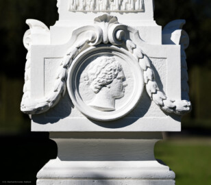 Schloss und Schlossgarten Schwetzingen - Garten - Kreisparterre, nordöstliches Rasenfeld, entlang der Nord-Süd-Achse - Nordöstlicher Obelisk, Südseite - Blick auf ein weibliches verwittertes Profilbildnis / Flachrelief, umgeben von einem Lorbeerfeston (Person nicht eindeutig identifizierbar) (Bildhauer: Peter Anton von Verschaffelt (1710-1793); Datierung: 1762-69; Obelisk Teil einer Vierergruppe im Kreisparterre; Material: roter Sandstein (Medaillons aus gelbem Sandstein); Fassung: Weiß; Obelisk dreigeteilt; Sockel, Block mit Medaillonbildnissen, eigentlicher Obelisk; Abmessungen: Höhe ca. 6 Meter, Sockel Höhe 155 cm / Breite 87 cm, Block mit Medaillons Höhe 67 cm / Breite 87 cm; Obelisk oben mit Tropfsteindekor) (aufgenommen im September 2023, am Nachmittag)