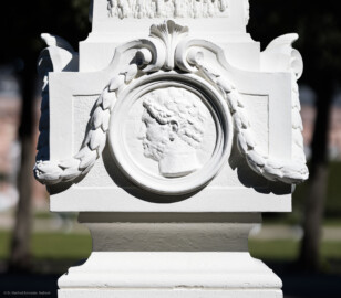 Schloss und Schlossgarten Schwetzingen - Garten - Kreisparterre, nordöstliches Rasenfeld, entlang der Nord-Süd-Achse - Nordöstlicher Obelisk, Westseite - Blick auf ein männliches verwittertes Profilbildnis / Flachrelief, umgeben von einem Lorbeerfeston (Person nicht eindeutig identifizierbar) (Bildhauer: Peter Anton von Verschaffelt (1710-1793); Datierung: 1762-69; Obelisk Teil einer Vierergruppe im Kreisparterre; Material: roter Sandstein (Medaillons aus gelbem Sandstein); Fassung: Weiß; Obelisk dreigeteilt; Sockel, Block mit Medaillonbildnissen, eigentlicher Obelisk; Abmessungen: Höhe ca. 6 Meter, Sockel Höhe 155 cm / Breite 87 cm, Block mit Medaillons Höhe 67 cm / Breite 87 cm; Obelisk oben mit Tropfsteindekor) (aufgenommen im September 2023, am Nachmittag) Schloss und Schlossgarten Schwetzingen - Garten - Kreisparterre, nordöstliches Rasenfeld, entlang der Nord-Süd-Achse - Nordöstlicher Obelisk, Westseite - Blick auf ein männliches verwittertes Profilbildnis / Flachrelief, umgeben von einem Lorbeerfeston (Person nicht eindeutig identifizierbar) (Bildhauer: Peter Anton von Verschaffelt (1710-1793); Datierung: 1762-69; Obelisk Teil einer Vierergruppe im Kreisparterre; Material: roter Sandstein (Medaillons aus gelbem Sandstein); Fassung: Weiß; Obelisk dreigeteilt; Sockel, Block mit Medaillonbildnissen, eigentlicher Obelisk; Abmessungen: Höhe ca. 6 Meter, Sockel Höhe 155 cm / Breite 87 cm, Block mit Medaillons Höhe 67 cm / Breite 87 cm; Obelisk oben mit Tropfsteindekor) (aufgenommen im September 2023, am Nachmittag)