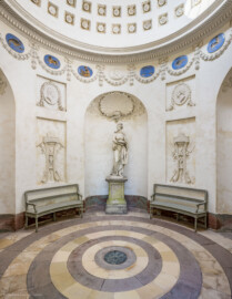 Schloss und Schlossgarten Schwetzingen - Garten - Tempel der Botanik - Von innen - Raum - Von Osten - Blick von der Eingangstür nach Westen in den Raum; in der Mitte in einer Rundbogennische die Skulptur der Botanik auf einem Postament (Bildhauer: Skulptur Francesco Carabelli (1737-1798), Postament Franz Conrad Linck (1730-1793), 1779); über der Skulptur eine Muschel, umgeben von zwei gekreuzten Eichenlaubzweigen und Blüten; links und rechts unten zwei Stuckreliefs mit Dreifuß, Feuerschale und Schlange, links mit zwei Harken und Blütenzweigen, den Frühling darstellend, rechts zwei gekreuzte Spaten mit Zweigen und Früchten, den Sommer darstellend; links und rechts oben zwei stuckierte Reliefporträts von Naturforschern, links Carl von Linné (1707-1778), rechts Joseph Pitton de Tournefort (1656-1708); unterhalb der Kuppel Girlanden, dazwischen Tierkreiszeichen und Stuckreliefs mit Putti, die Jahreszeiten darstellend; in der Kuppel rosettengeschmückte kleine Kassetten; auf dem Fußboden konzentrische Sandsteinplatten, ähnlich Jahresringen eines Baumes, in der Mitte eine kreisförmige Öffnung, bedeckt mit einem Blech, zum Blick in den dunklen, gewölbten, nicht zugänglichen Kellerraum (Tempel: Durchmesser ca. 7 Meter; Bauzeit: 1777-1780; Architekt: Nicolas de Pigage) (aufgenommen im September 2023, am frühen Nachmittag)