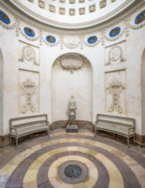 Schloss und Schlossgarten Schwetzingen - Garten - Tempel der Botanik - Von innen - Raum - Von Norden - Blick nach Süden in den Raum; in der Mitte in einer Rundbogennische eine Vase mit Postament (Bildhauer Postament: Franz Conrad Linck (1730-1793), um 1779; Bildhauer Vase: unklar, evtl. Peter Anton von Verschaffelt (1710-1793), Francesco Carabelli (1737-1798) oder Franz Conrad Linck (1730-1793)); oberhalb der Vase eine Muschel, umgeben von zwei gekreuzten Eichenlaubzweigen und Blüten; links und rechts unten zwei Stuckreliefs mit Dreifuß, Feuerschale und Schlange, links mit zwei gekreuzten Thyrsosstäben mit Weinranken und Trauben, den Herbst darstellend, rechts zwei Harken mit Blütenzweigen, den Frühling darstellend; links und rechts oben zwei stuckierte Reliefporträts von Naturforschern, links Theophrastos von Eresos (371-287 v. Chr.), rechts Carl von Linné (1707-1778); unterhalb der Kuppel Girlanden, dazwischen Tierkreiszeichen und Stuckreliefs mit Putti, die Jahreszeiten darstellend; in der Kuppel rosettengeschmückte kleine Kassetten; auf dem Fußboden konzentrische Sandsteinplatten, ähnlich Jahresringen eines Baumes, in der Mitte eine kreisförmige Öffnung, bedeckt mit einem Blech, darunter ein dunkler, gewölbter, nicht zugänglicher Kellerraum (Tempel: Durchmesser ca. 7 Meter; Bauzeit: 1777-1780; Architekt: Nicolas de Pigage) (aufgenommen im September 2023, am frühen Nachmittag)