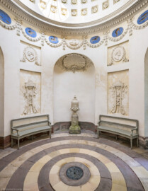 Schloss und Schlossgarten Schwetzingen - Garten - Tempel der Botanik - Von innen - Raum - Von Süden - Blick nach Norden in den Raum; in der Mitte in einer Rundbogennische eine Vase mit Postament (Bildhauer Postament: Franz Conrad Linck (1730-1793), um 1779; Bildhauer Vase: unklar, evtl. Peter Anton von Verschaffelt (1710-1793), Francesco Carabelli (1737-1798) oder Franz Conrad Linck (1730-1793)); oberhalb der Vase eine Muschel, umgeben von zwei gekreuzten Eichenlaubzweigen und Blüten; links und rechts unten zwei Stuckreliefs mit Dreifuß, Feuerschale und Schlange, links mit zwei gekreuzten Spaten mit Zweigen und Früchten, den Sommer darstellend, rechts zwei gekreuzte Krummstäbe (wohl Schäferstäbe) mit Eichenlaub, den Winter darstellend; links und rechts oben zwei stuckierte Reliefporträts von Naturforschern, links Joseph Pitton de Tournefort (1656-1708), rechts Plinius der Ältere (23-79); unterhalb der Kuppel Girlanden, dazwischen Tierkreiszeichen und Stuckreliefs mit Putti, die Jahreszeiten darstellend; in der Kuppel rosettengeschmückte kleine Kassetten; auf dem Fußboden konzentrische Sandsteinplatten, ähnlich Jahresringen eines Baumes, in der Mitte eine kreisförmige Öffnung, bedeckt mit einem Blech, darunter ein dunkler, gewölbter, nicht zugänglicher Kellerraum (Tempel: Durchmesser ca. 7 Meter; Bauzeit: 1777-1780; Architekt: Nicolas de Pigage) (aufgenommen im September 2023, am frühen Nachmittag)